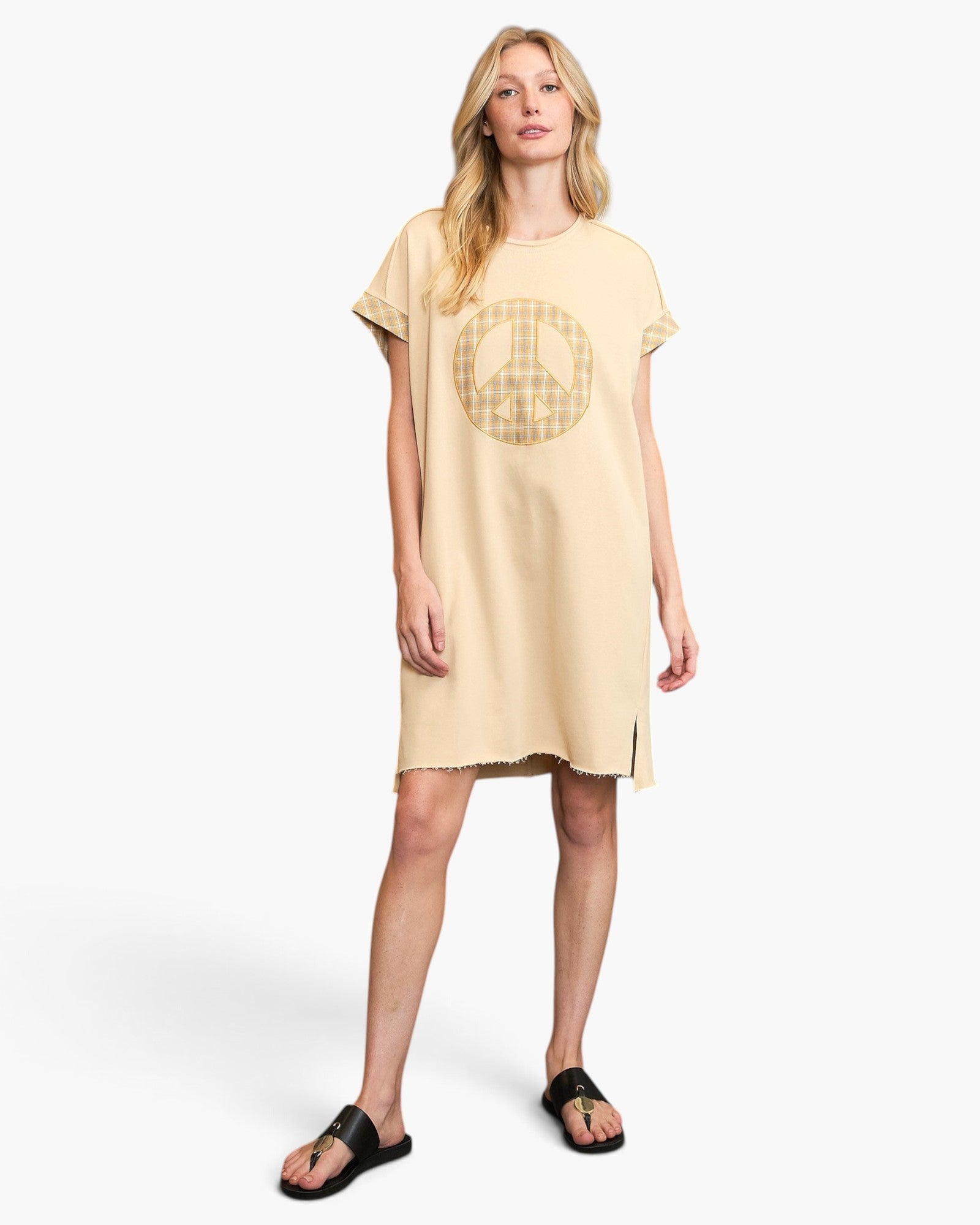 A woman modeling a beige short-sleeve tunic dress with a gold peace sign graphic on the front, paired with black slide sandals, against a light gray studio background.