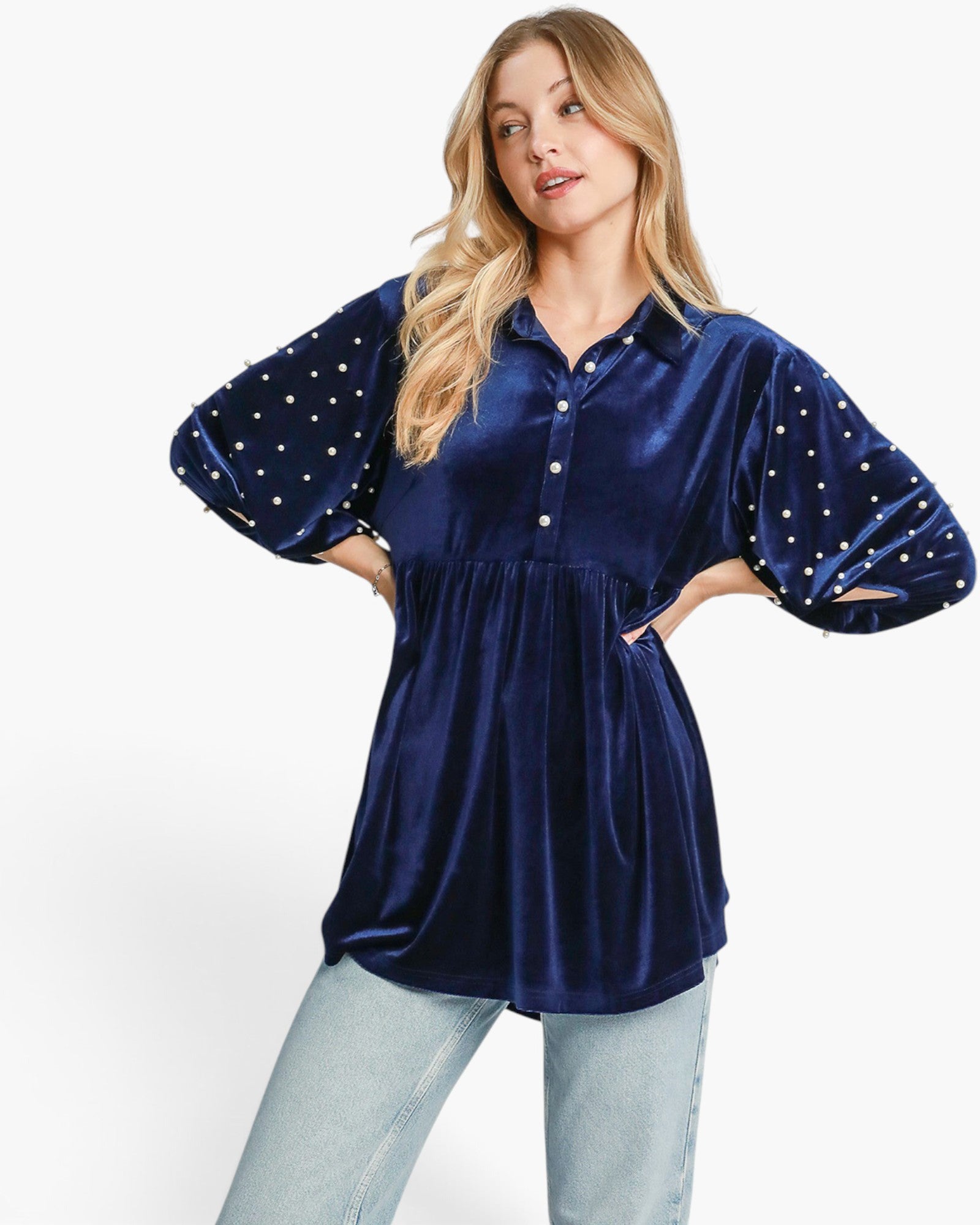 A woman modeling a navy blue velvet button-up tunic top with white polka-dot patterned balloon sleeves, paired with light-wash jeans, against a plain white studio background.