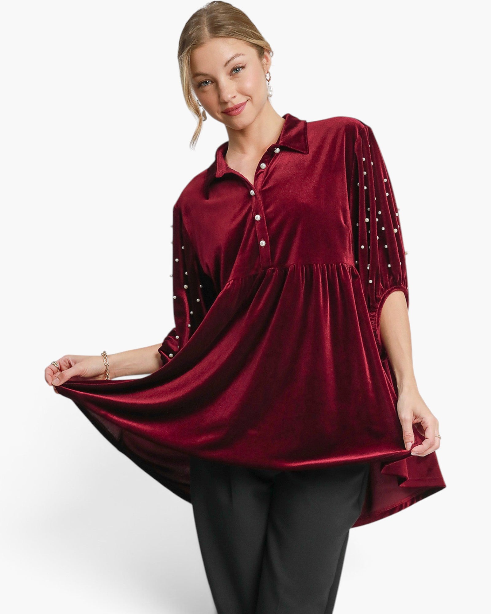 A woman modeling a deep burgundy velvet tunic top with three-quarter sleeves, adorned with small pearl-like embellishments along the sleeve edges, paired with black pants against a clean white studio background.