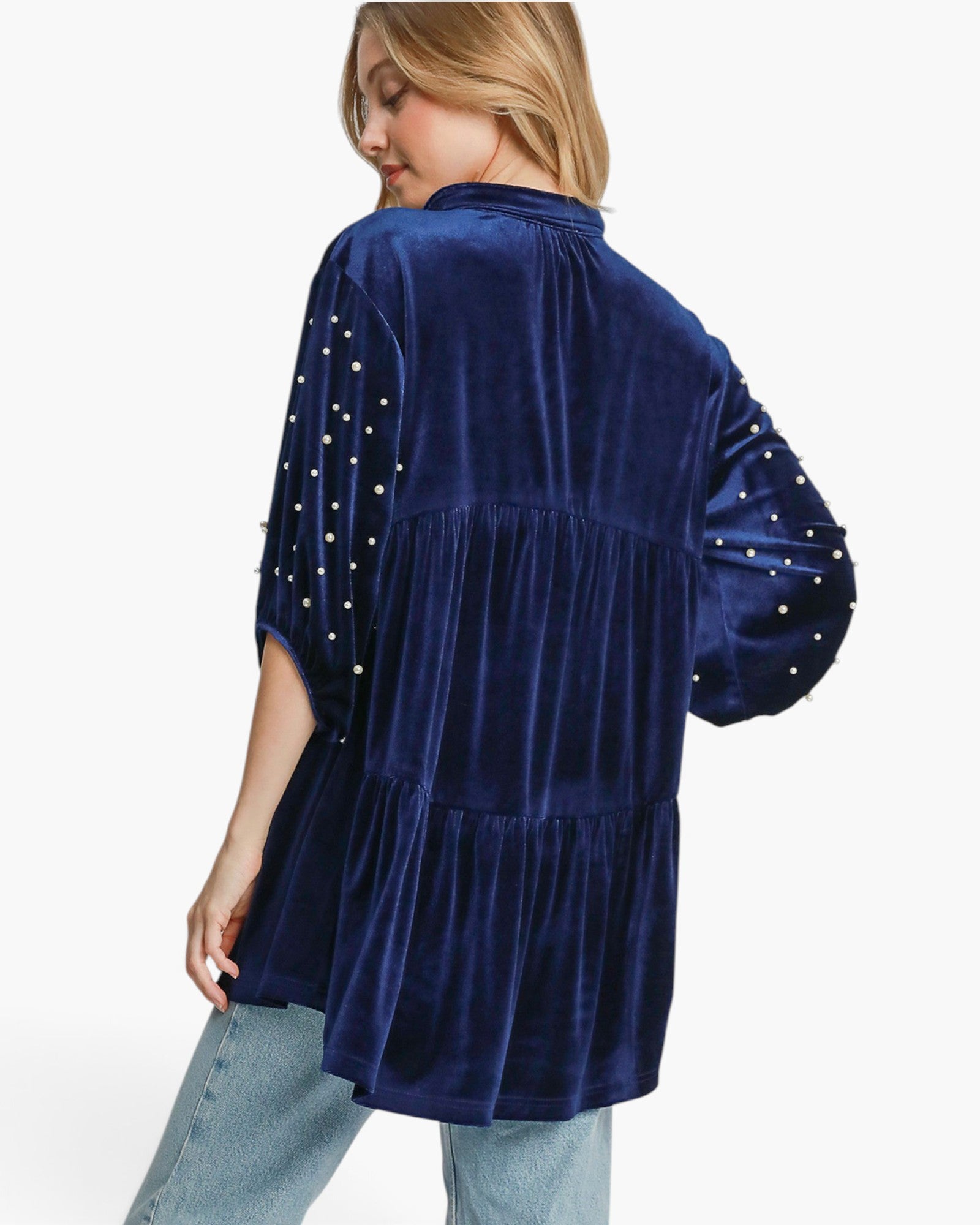 A woman wearing a navy blue velvet tiered blouse with pearl embellishments on the sleeves, shown from a three-quarter back angle against a plain white background.