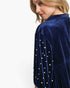 A close-up side view of a woman wearing a navy blue velvet top with pearl embellishments on the sleeve, against a plain white background.