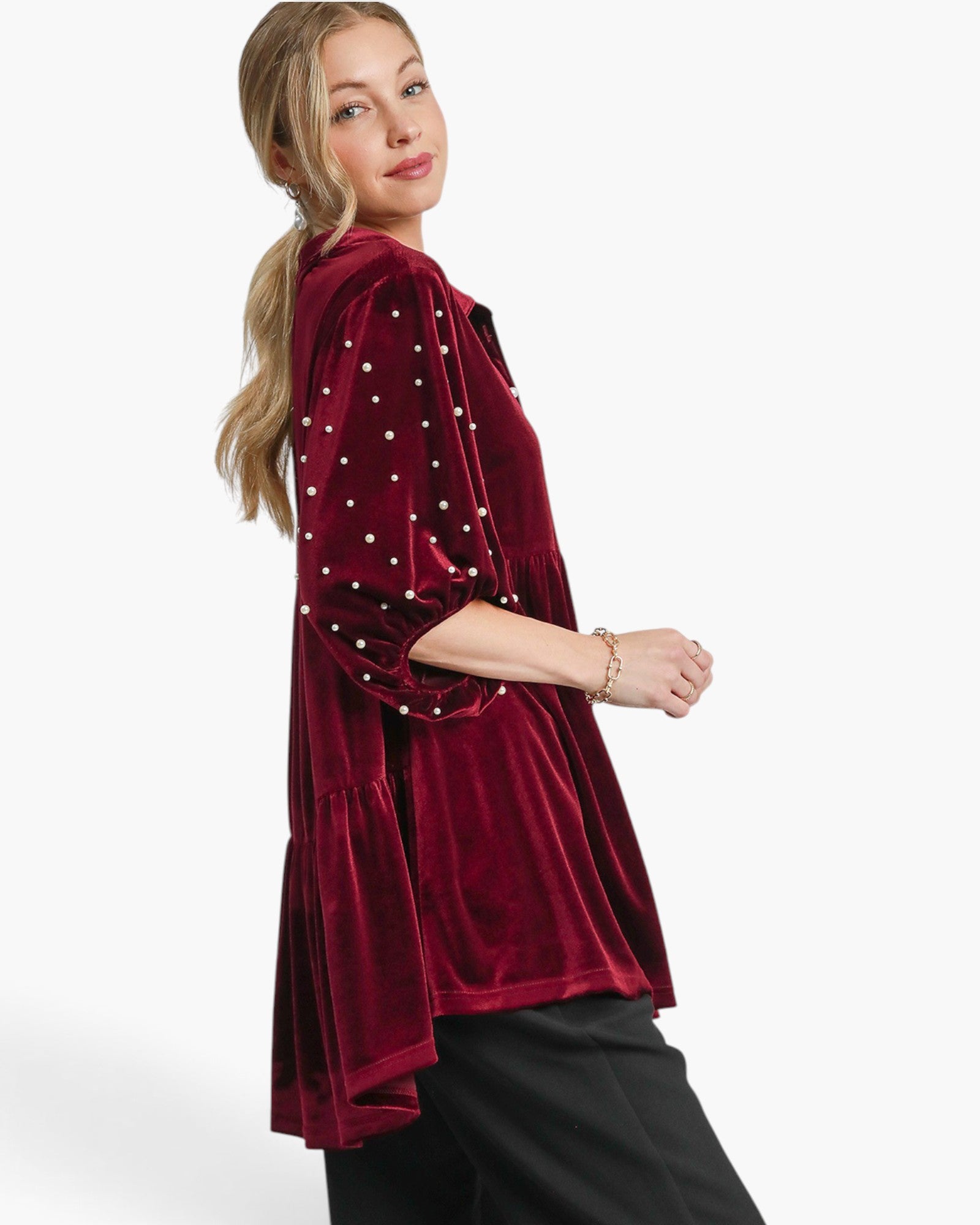 A woman wearing a deep burgundy velvet tunic top with white embroidered dots and black pants, photographed against a clean white studio background.