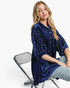 A woman wearing a navy blue velvet button-up shirt with white pearl embellishments, paired with light blue jeans, sitting on a modern folding chair against a white studio background.