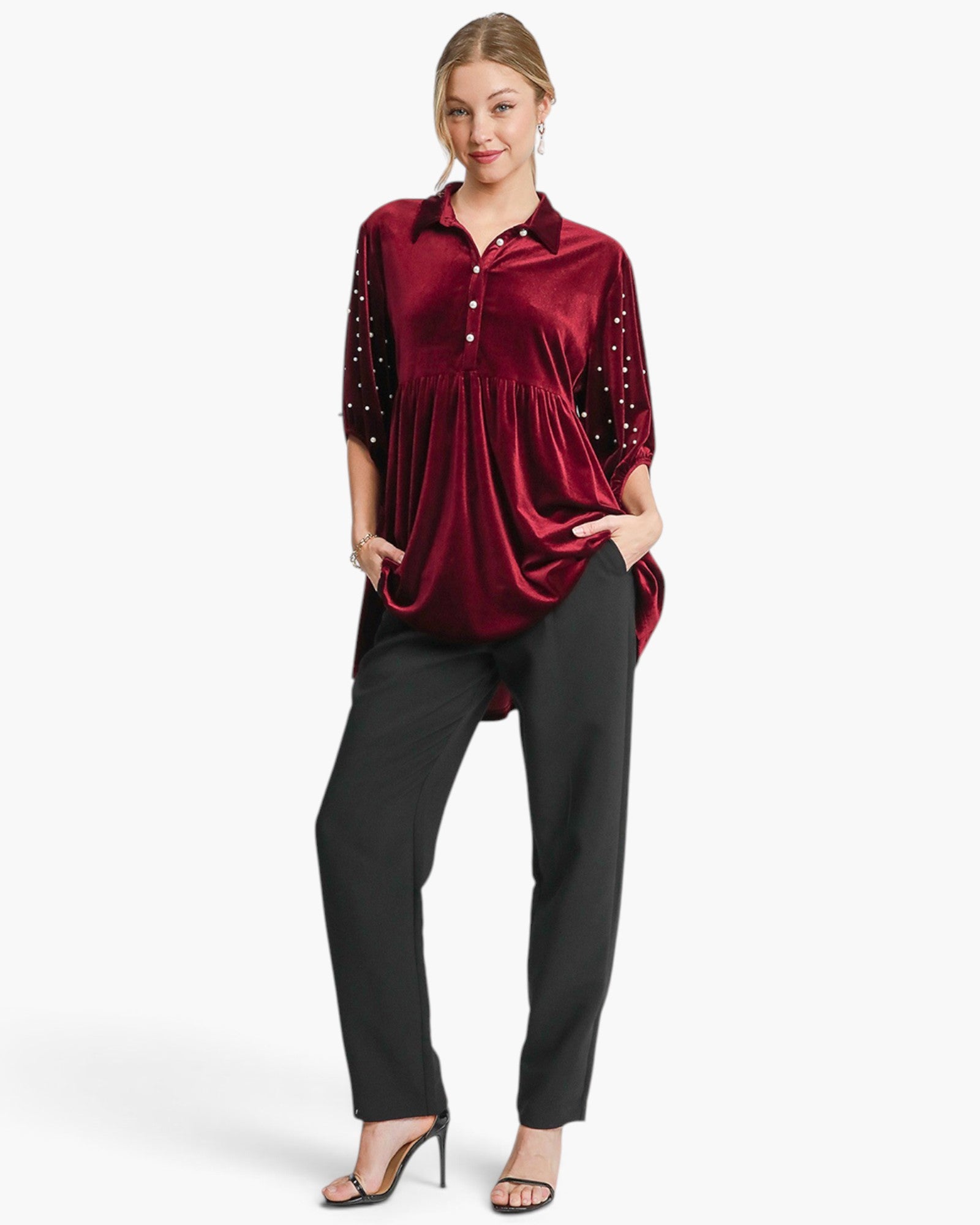 A woman modeling a burgundy velvet long-sleeve top with silver rhinestone embellishments on the sleeves, paired with black pants and black heeled sandals, against a light gray studio background.