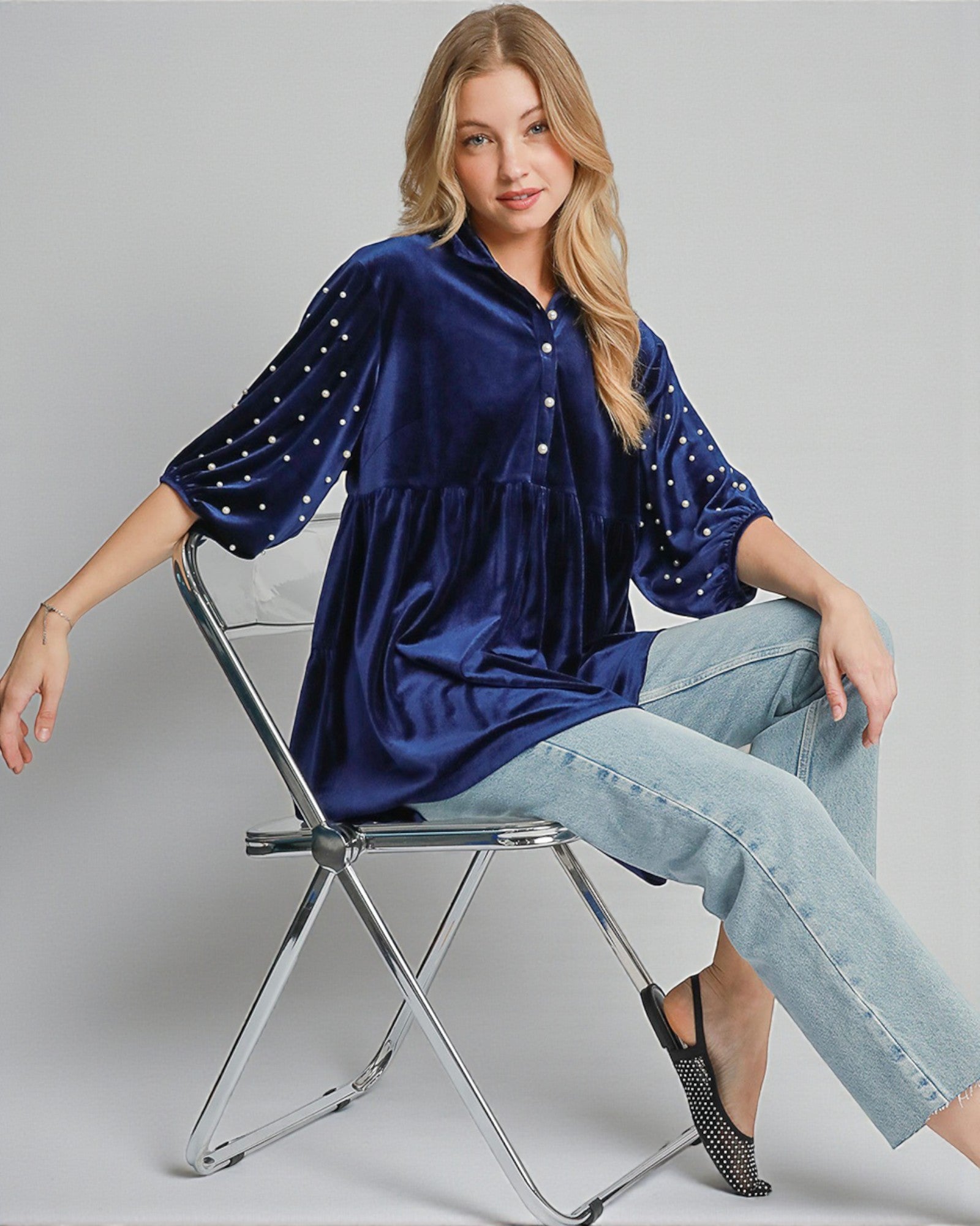 Woman wearing a blue dress with white embellishments sitting on a chair against a gray background