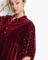 A woman wearing a deep burgundy velvet shirt dress with pearl embellishments on the sleeves and collar, photographed against a clean white background.