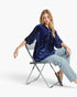 A woman sits on a folding chair wearing a navy blue velvet tunic top with silver rhinestone embellishments on the sleeves, paired with light-wash cropped jeans and black pointed flats, against a plain white studio background.