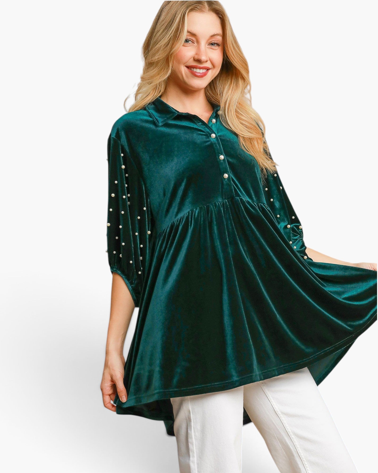A woman wearing a dark green velvet tunic top with pearl embellishments on the sleeves, paired with white pants, against a plain white background.