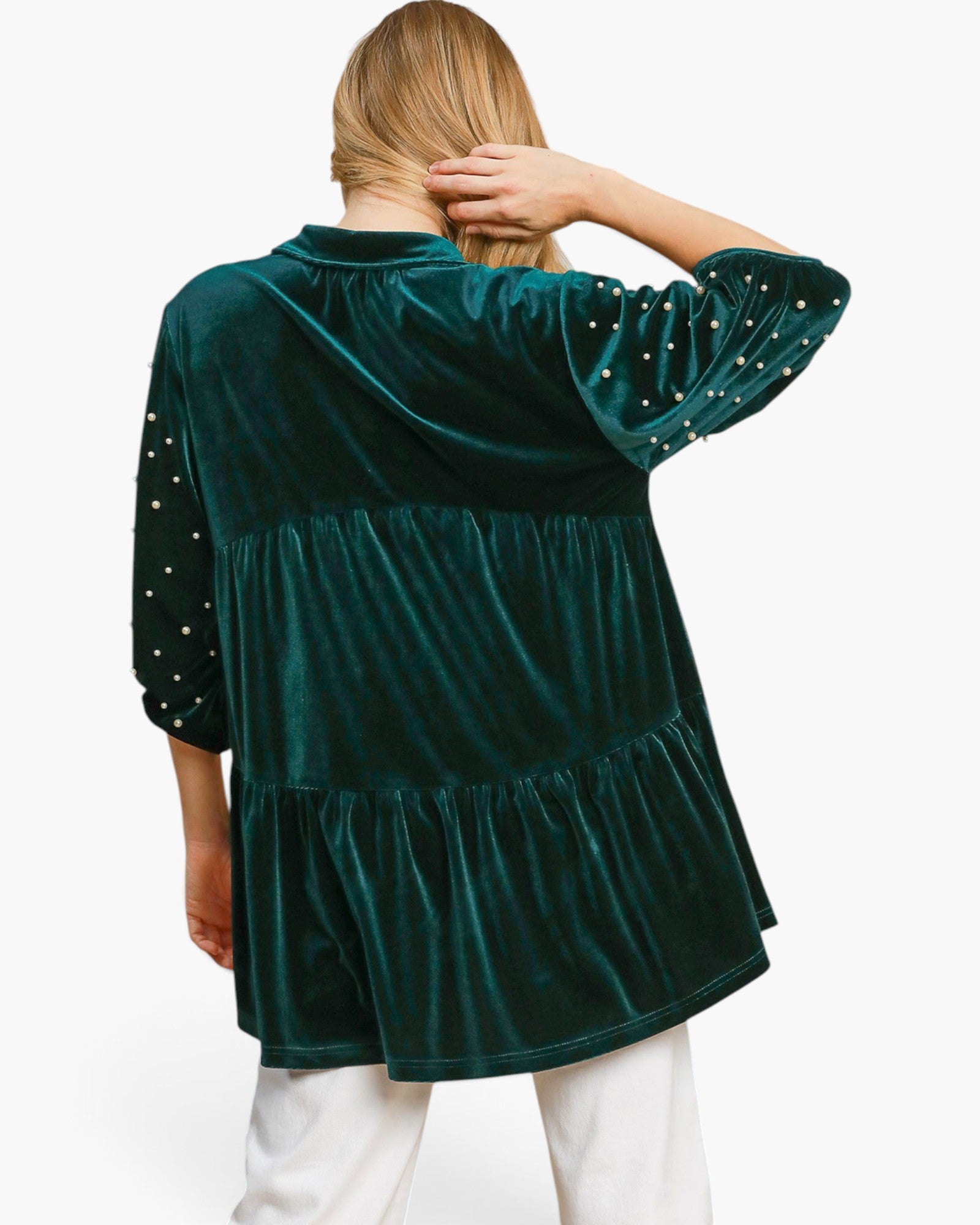 A woman shown from the back wearing a dark green velvet tiered tunic top with dotted sleeves, paired with white pants, against a plain white background.