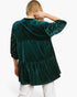 A woman shown from the back wearing a dark green velvet tiered tunic top with dotted sleeves, paired with white pants, against a plain white background.