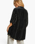 A woman shown from the back wearing a black velvet tiered tunic top with embellished sleeves, paired with light gray pants, against a plain white studio background.