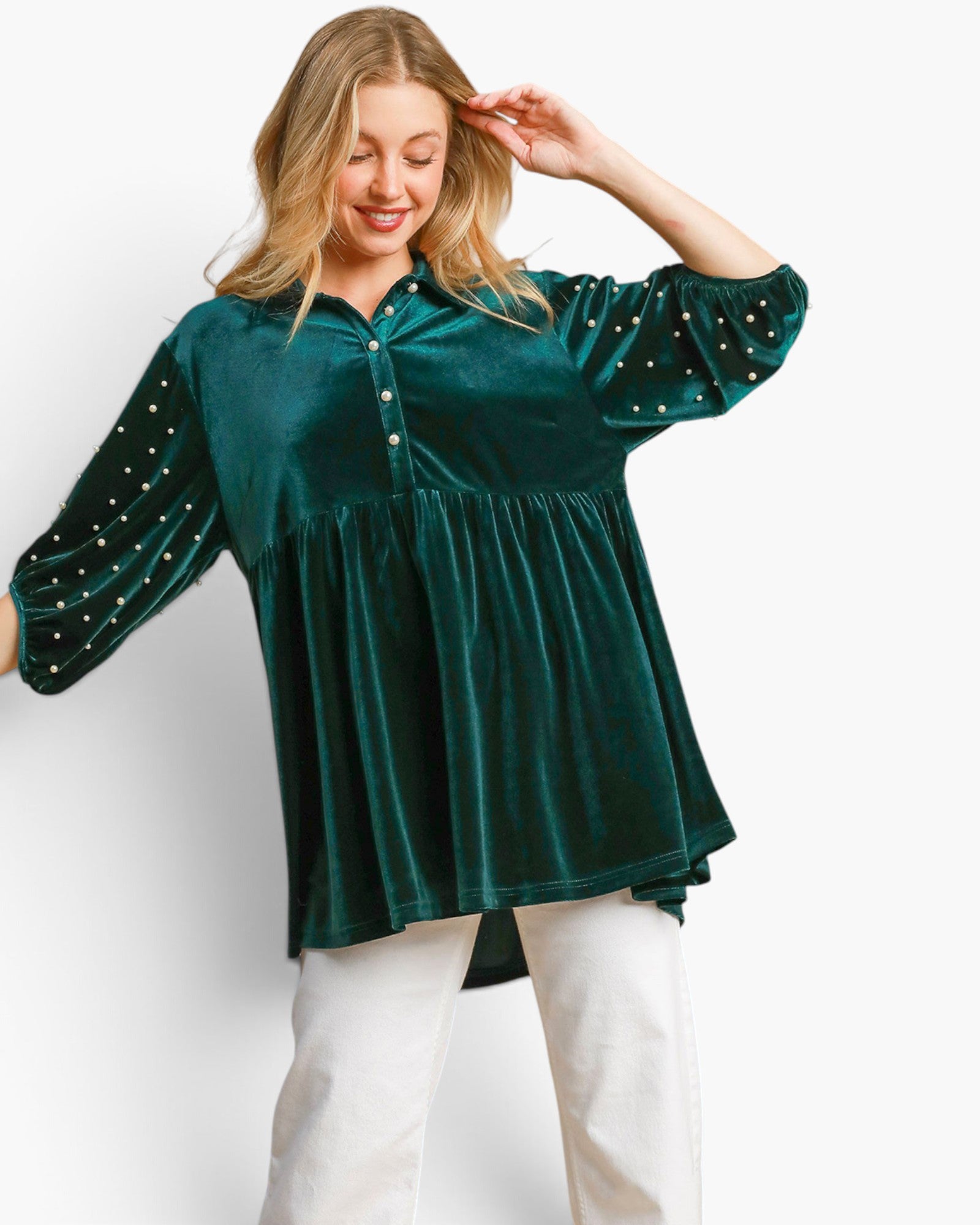 A woman wearing a dark green velvet long-sleeve top with white beaded trim on the sleeves, paired with white pants, against a plain white background.