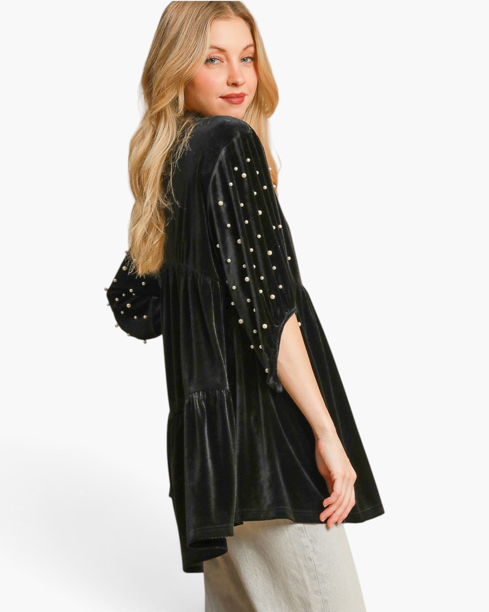 A woman modeling a black velvet long-sleeve top with gold beaded details on the sleeves, paired with light beige pants, against a plain white studio background.
