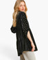A woman modeling a black velvet long-sleeve top with gold beaded details on the sleeves, paired with light beige pants, against a plain white studio background.