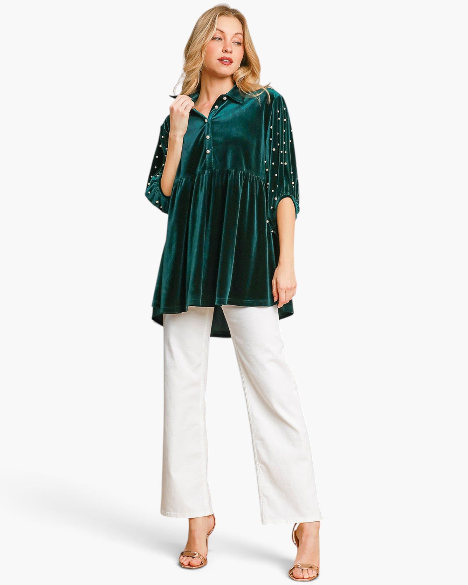 A woman modeling a dark green velvet tunic top with silver beaded fringe on the sleeves, paired with white wide-leg pants and clear high-heeled sandals, against a plain white background.