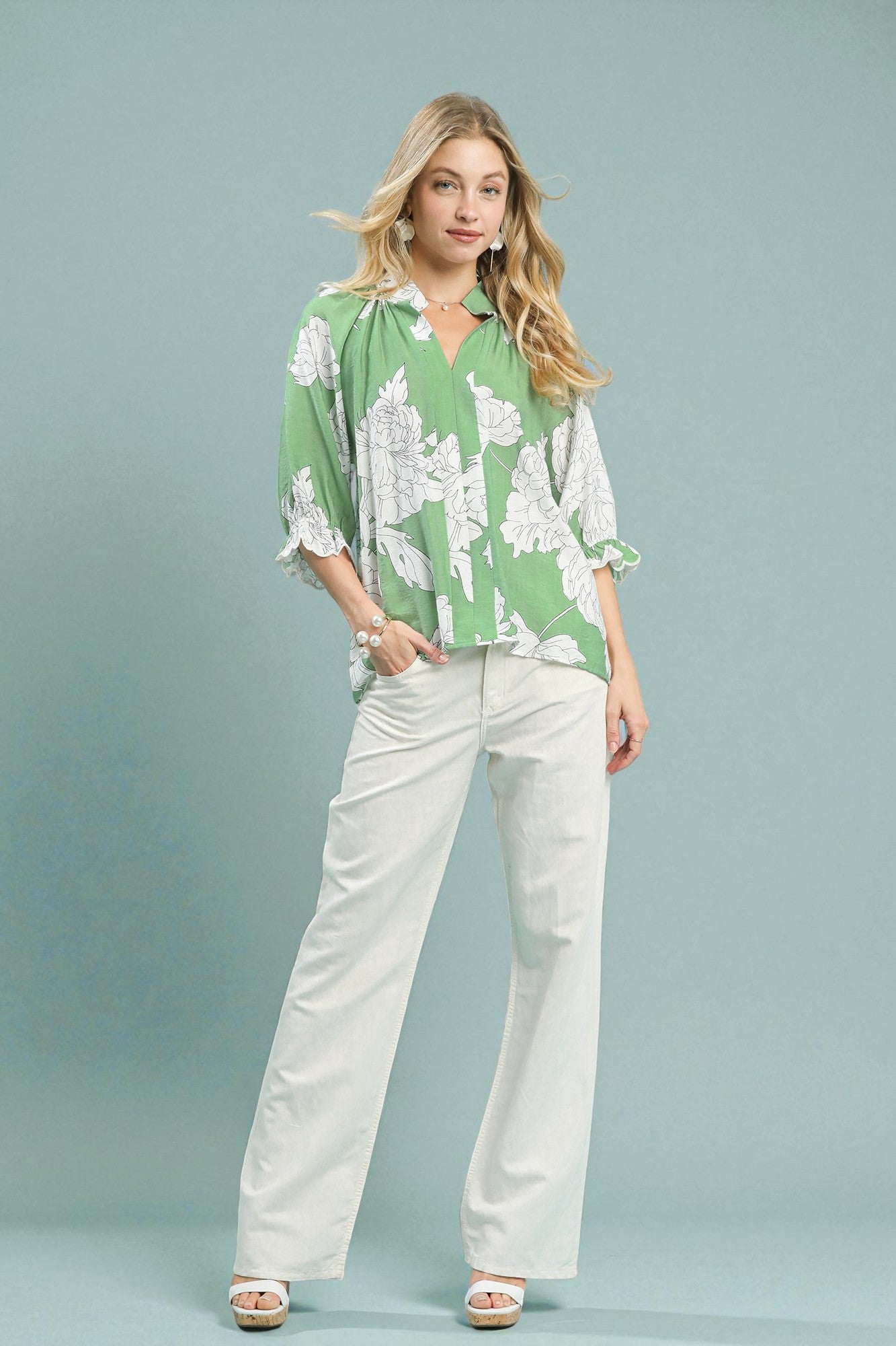 Woman wearing a green floral blouse and white pants against a gray background