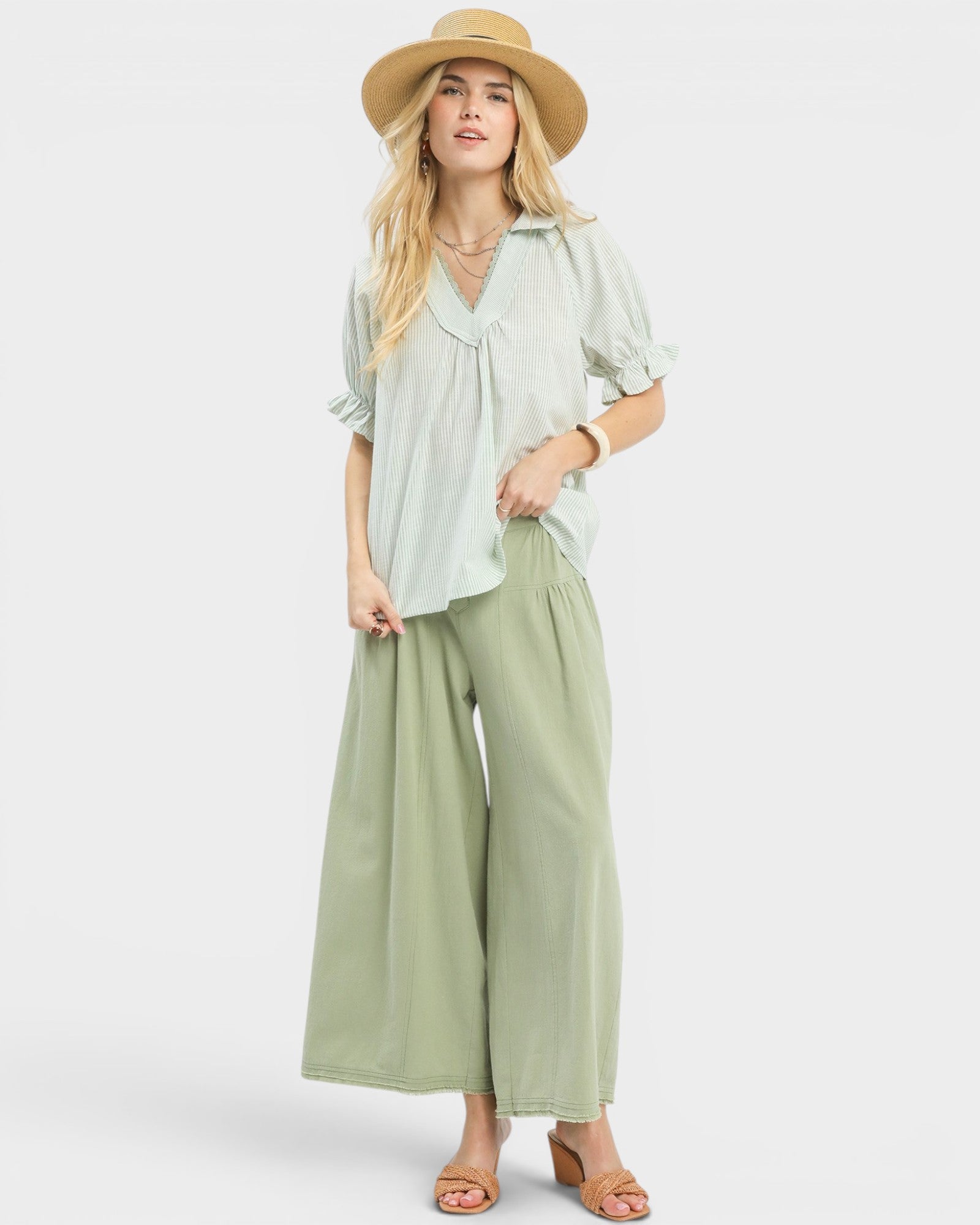Woman wearing a light green blouse and wide-leg pants with a straw hat on a white background