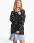 A woman modeling a black knit cardigan with white polka dots over a light blue collared shirt and a light blue frayed hem dress, set against a plain white studio background.