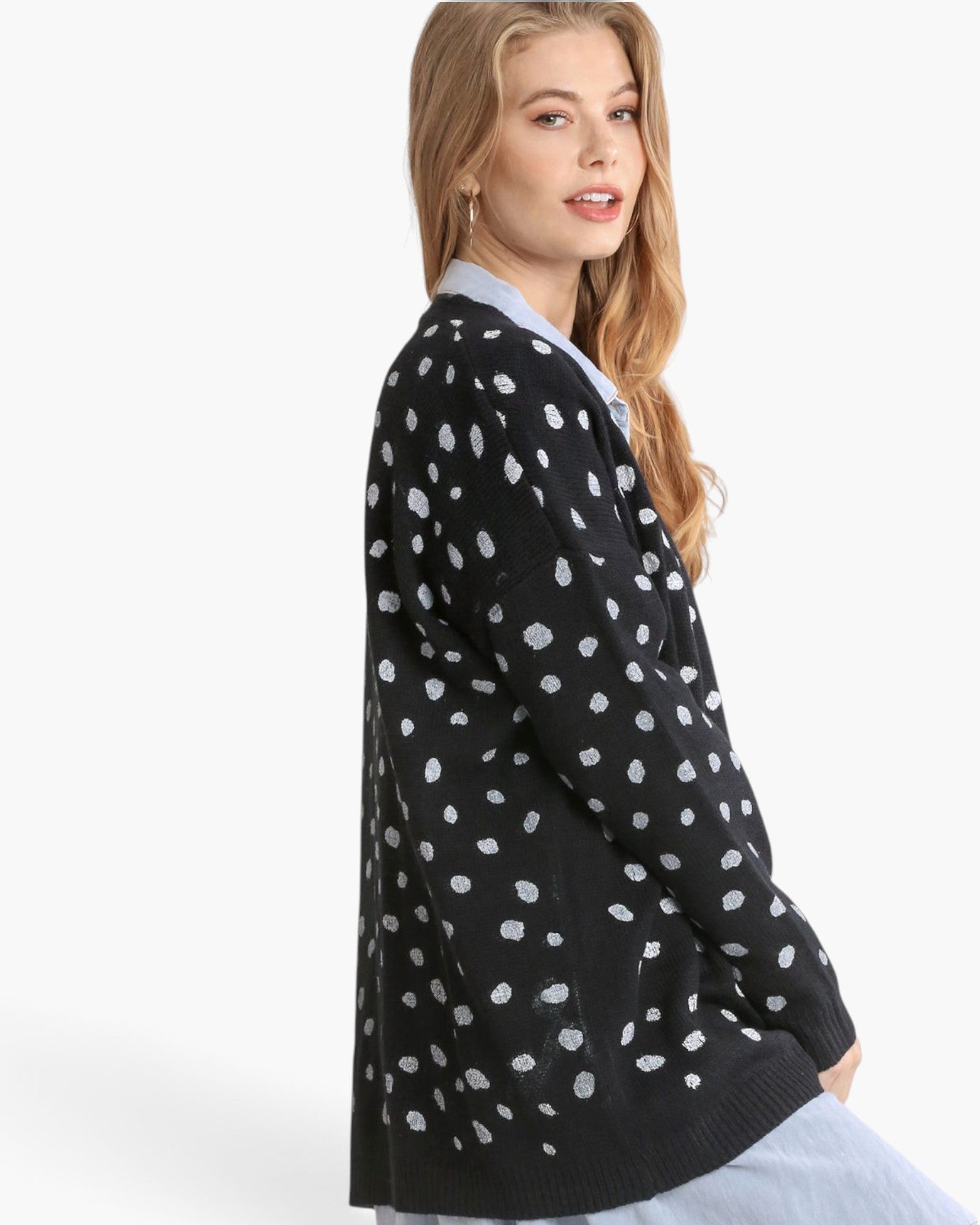 A woman wearing a black knit cardigan with a white spotted pattern, layered over a light blue collared shirt, shown from a side-back angle against a plain white studio background.