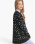 A woman wearing a black knit cardigan with a white spotted pattern, layered over a light blue collared shirt, shown from a side-back angle against a plain white studio background.
