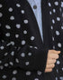 Close-up of a person wearing a black knit cardigan with white polka dots over a light blue button-up shirt. The cardigan has ribbed cuffs and a dark button closure.
