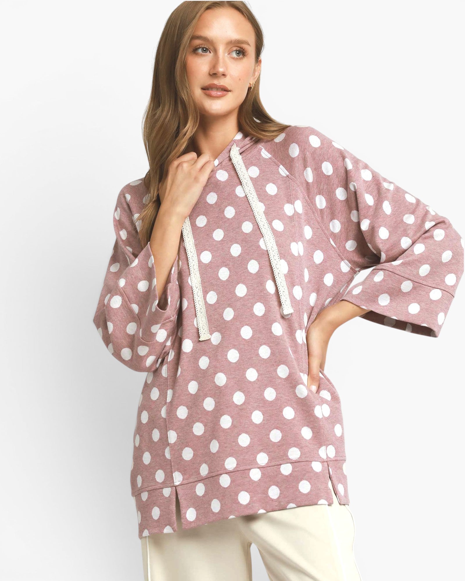 A woman wearing a dusty rose pink pullover hoodie with white polka dots and a drawstring hood, paired with cream pants, against a plain light gray background.