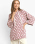 A woman wearing a dusty rose pink pullover hoodie with white polka dots and a drawstring hood, paired with cream pants, against a plain light gray background.
