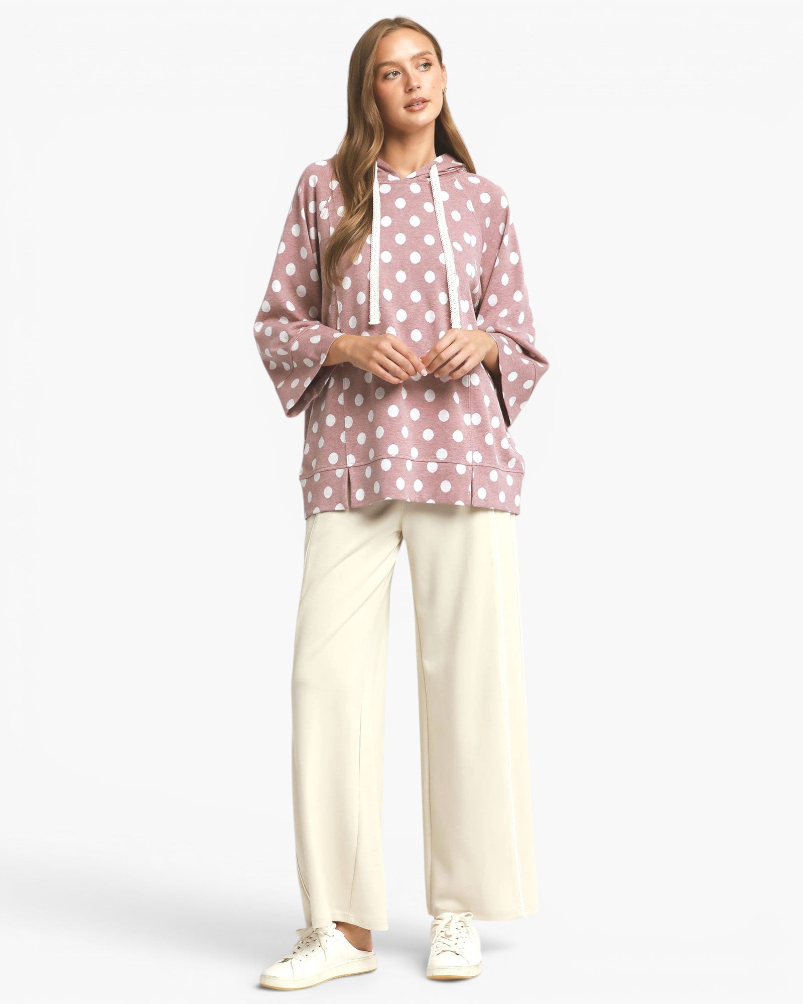 A woman modeling a dusty pink hooded sweatshirt with white polka dots and white drawstrings, paired with cream wide-leg pants and white sneakers, against a light gray studio background.