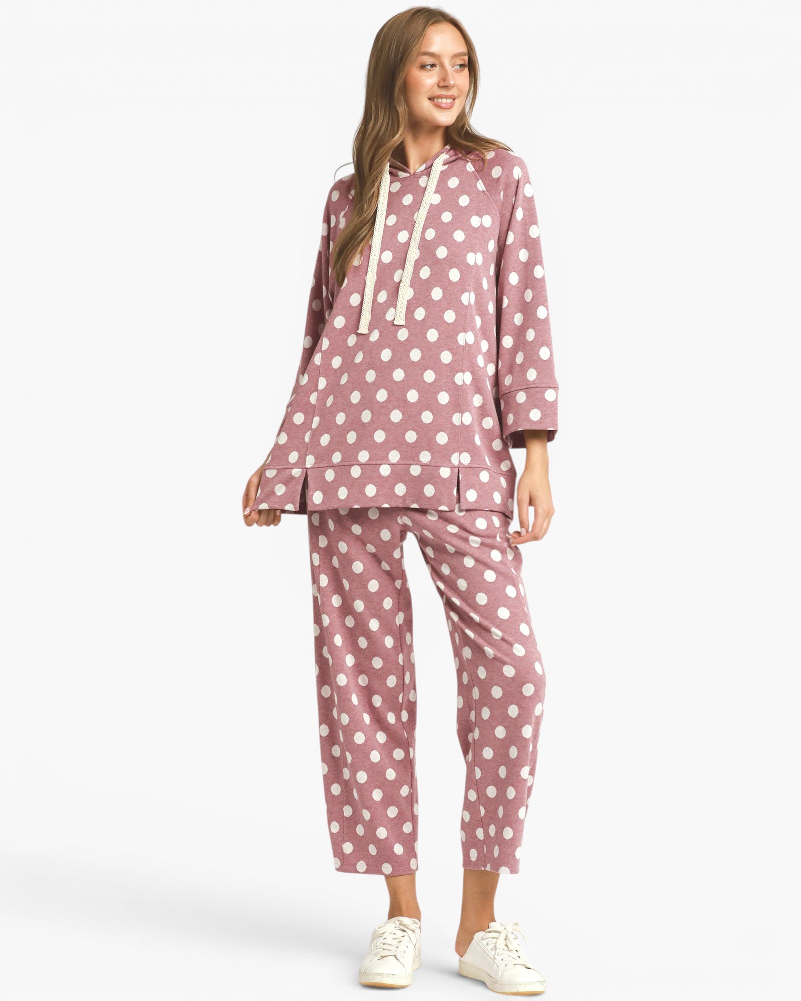 A woman modeling a two-piece pajama set featuring a dusty rose pink and white polka dot pattern, consisting of a hooded sweatshirt with white drawstrings and matching cropped pants, paired with white sneakers against a light gray background.
