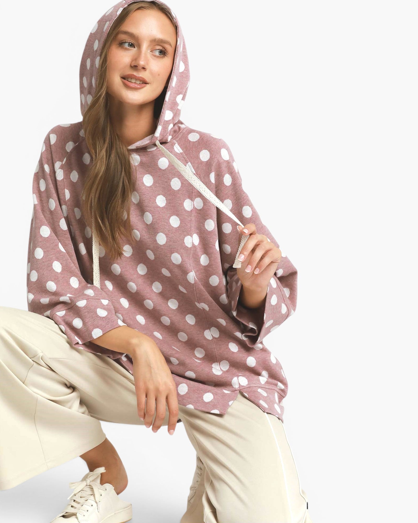 A woman wearing a dusty rose pink pullover hoodie with a white polka dot pattern, paired with beige pants and white sneakers, posing against a light gray studio background.