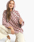 A woman wearing a dusty rose pink pullover hoodie with a white polka dot pattern, paired with beige pants and white sneakers, posing against a light gray studio background.