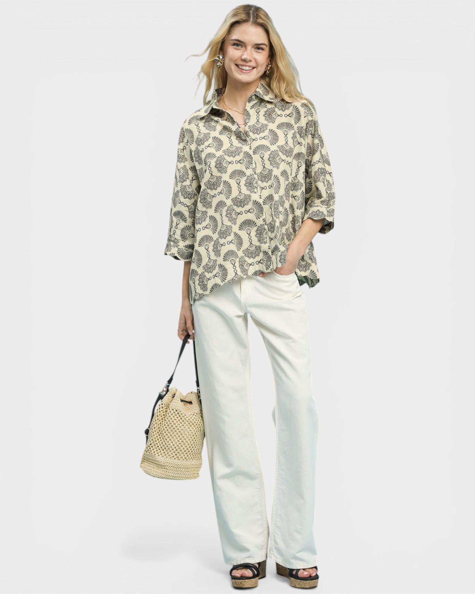 Woman wearing a patterned blouse and white pants, holding a straw bag on a plain background