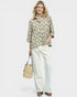 Woman wearing a patterned blouse and white pants, holding a straw bag on a plain background