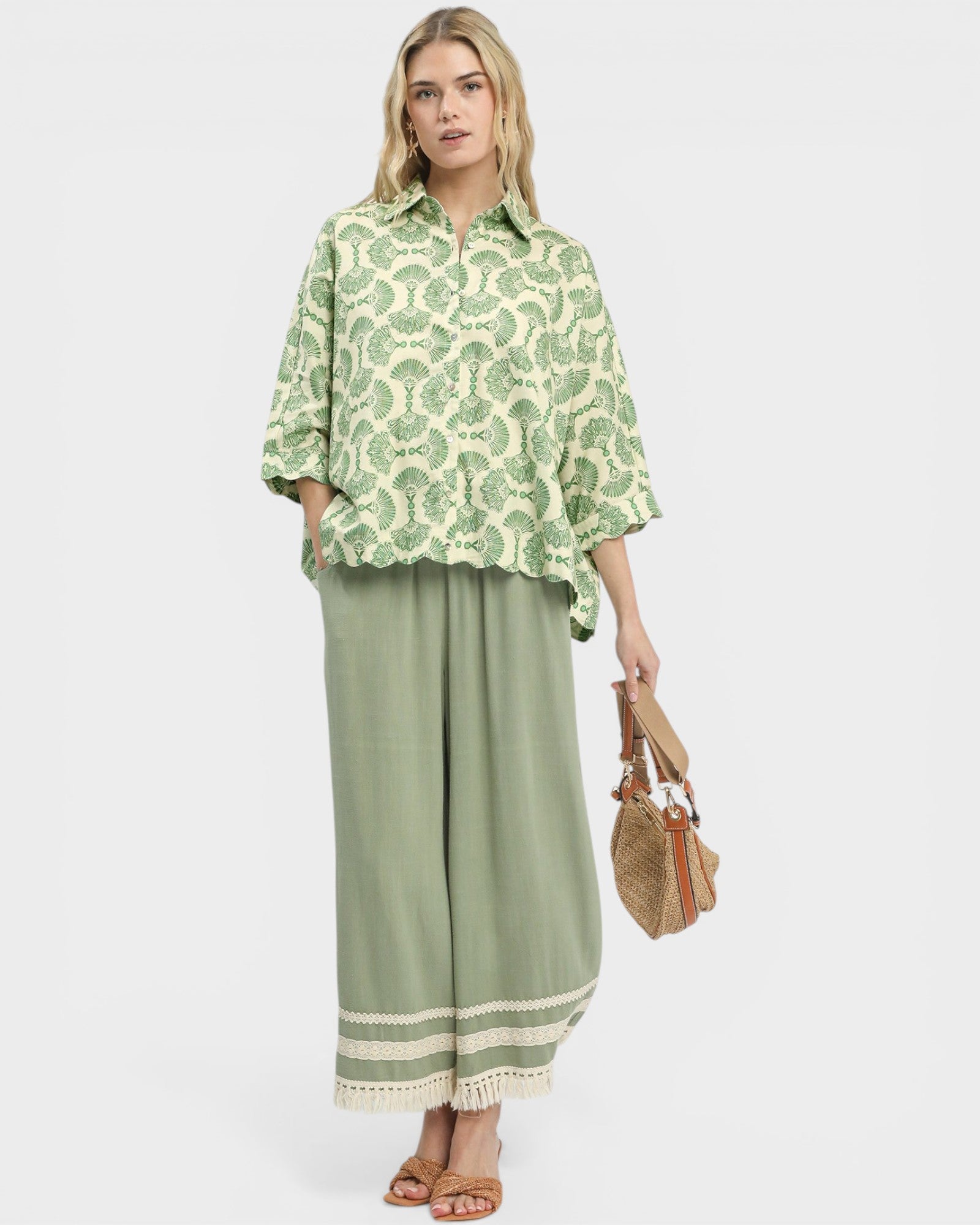 Woman wearing a green patterned blouse and green pants with a white border, holding a straw bag on a white background.