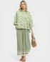 Woman wearing a green patterned blouse and green pants with a white border, holding a straw bag on a white background.
