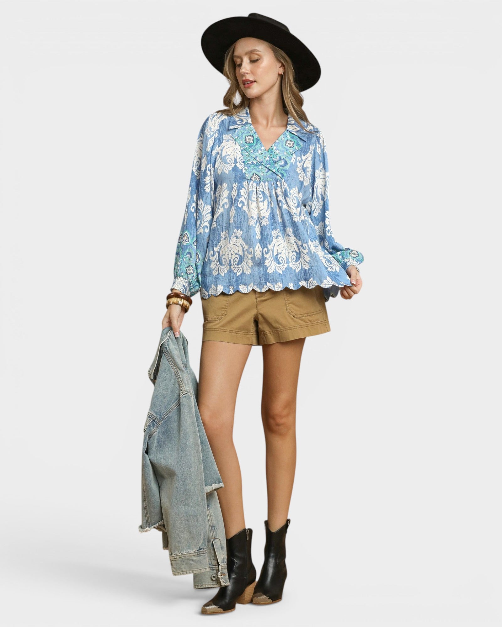 Woman wearing a blue floral blouse, beige shorts, black boots, and a black hat on a white background.