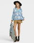 Woman wearing a blue floral blouse, beige shorts, black boots, and a black hat on a white background.
