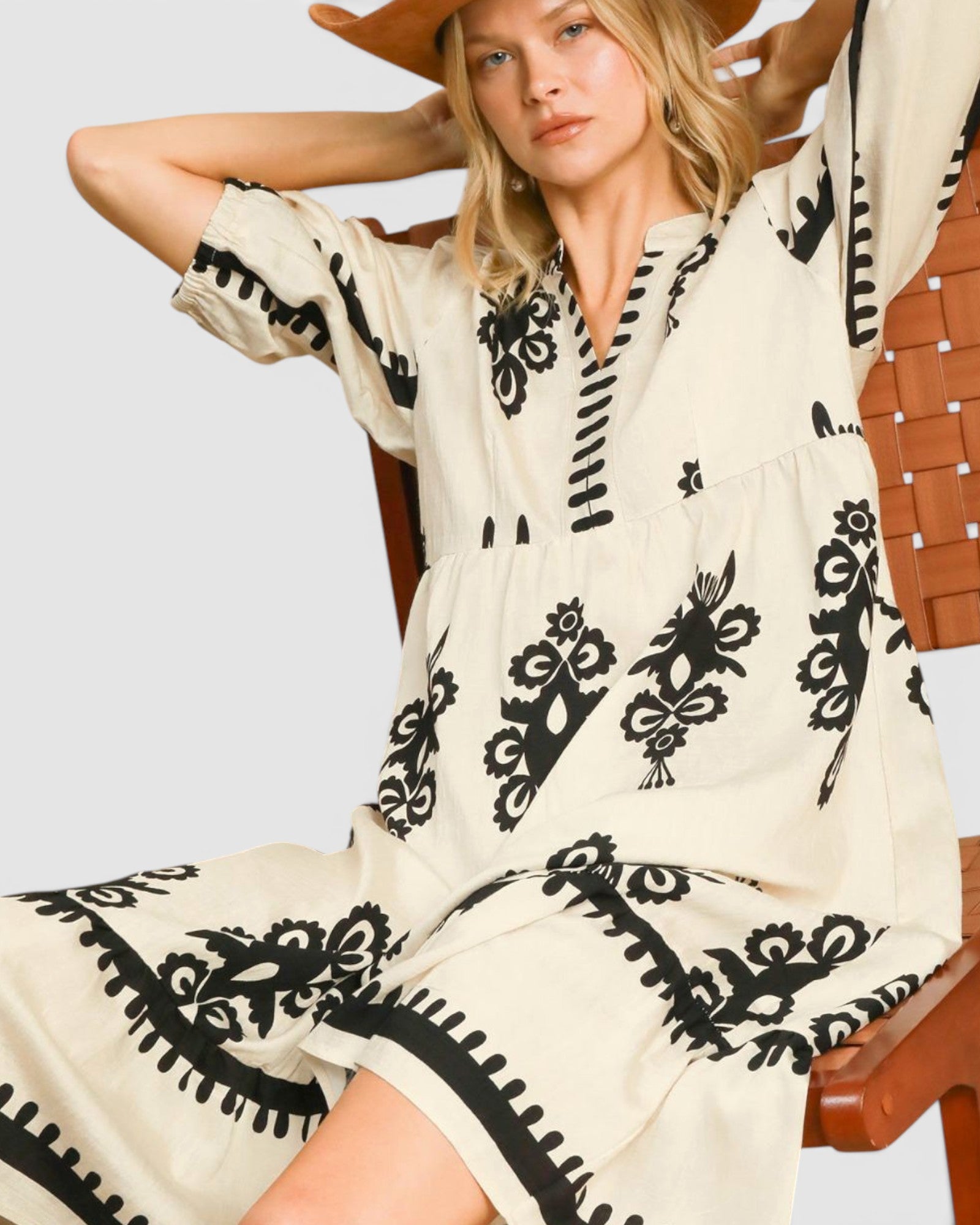 Woman wearing a white dress with black patterns sitting on a wooden chair.