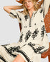 Woman wearing a white dress with black patterns sitting on a wooden chair.