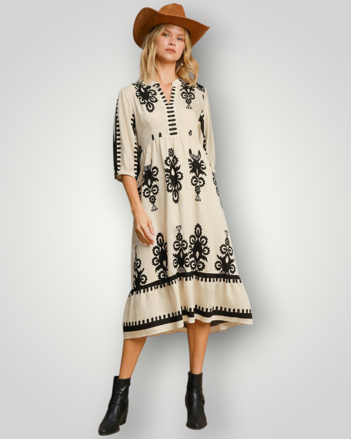 Woman wearing a cream dress with black patterns, black boots, and a brown hat on a white background
