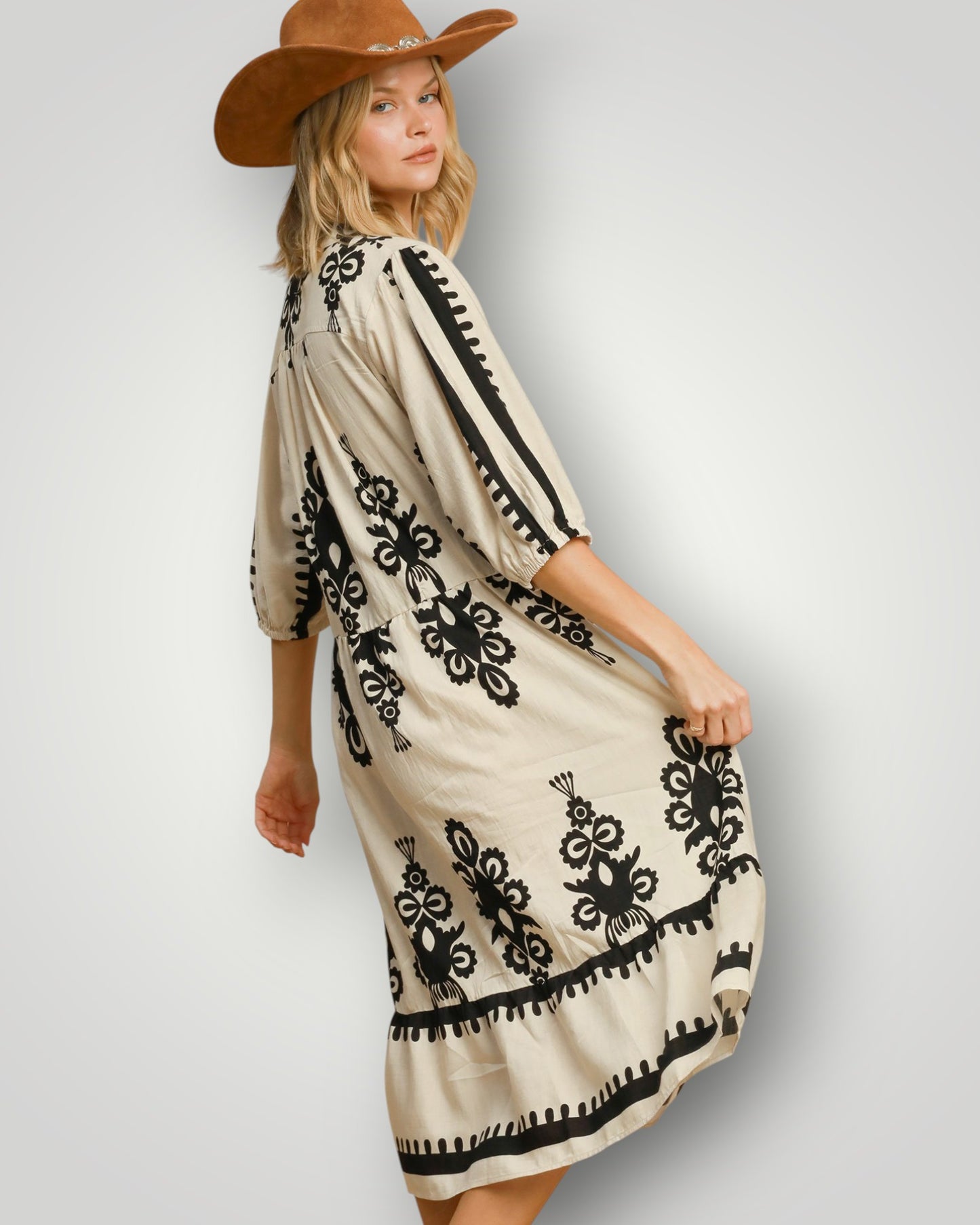 Woman wearing a beige dress with black embroidery and a brown cowboy hat on a light gray background