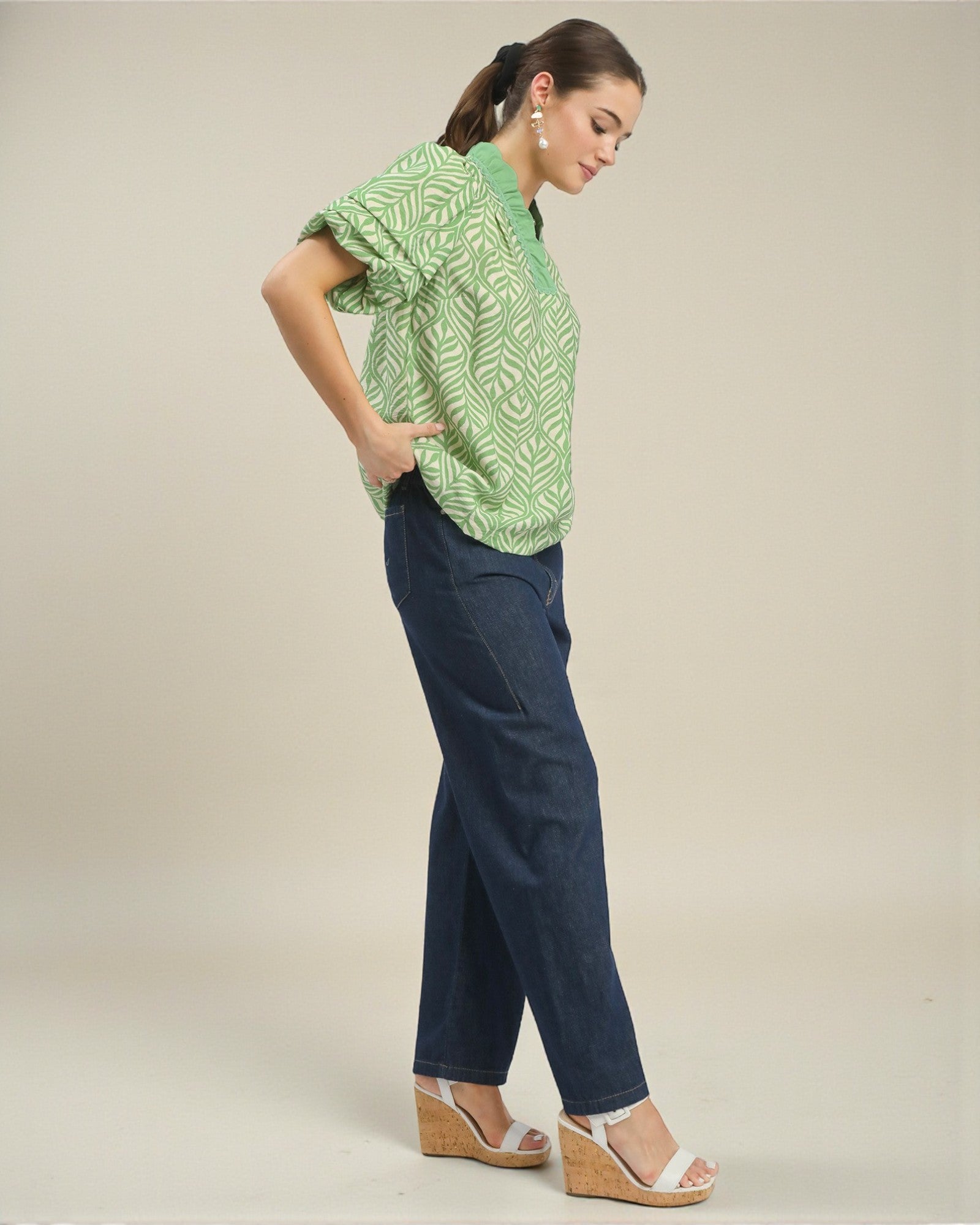 Woman wearing a green patterned top and navy pants on a beige background