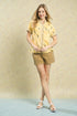 Woman wearing a yellow patterned shirt and beige shorts against a plain background