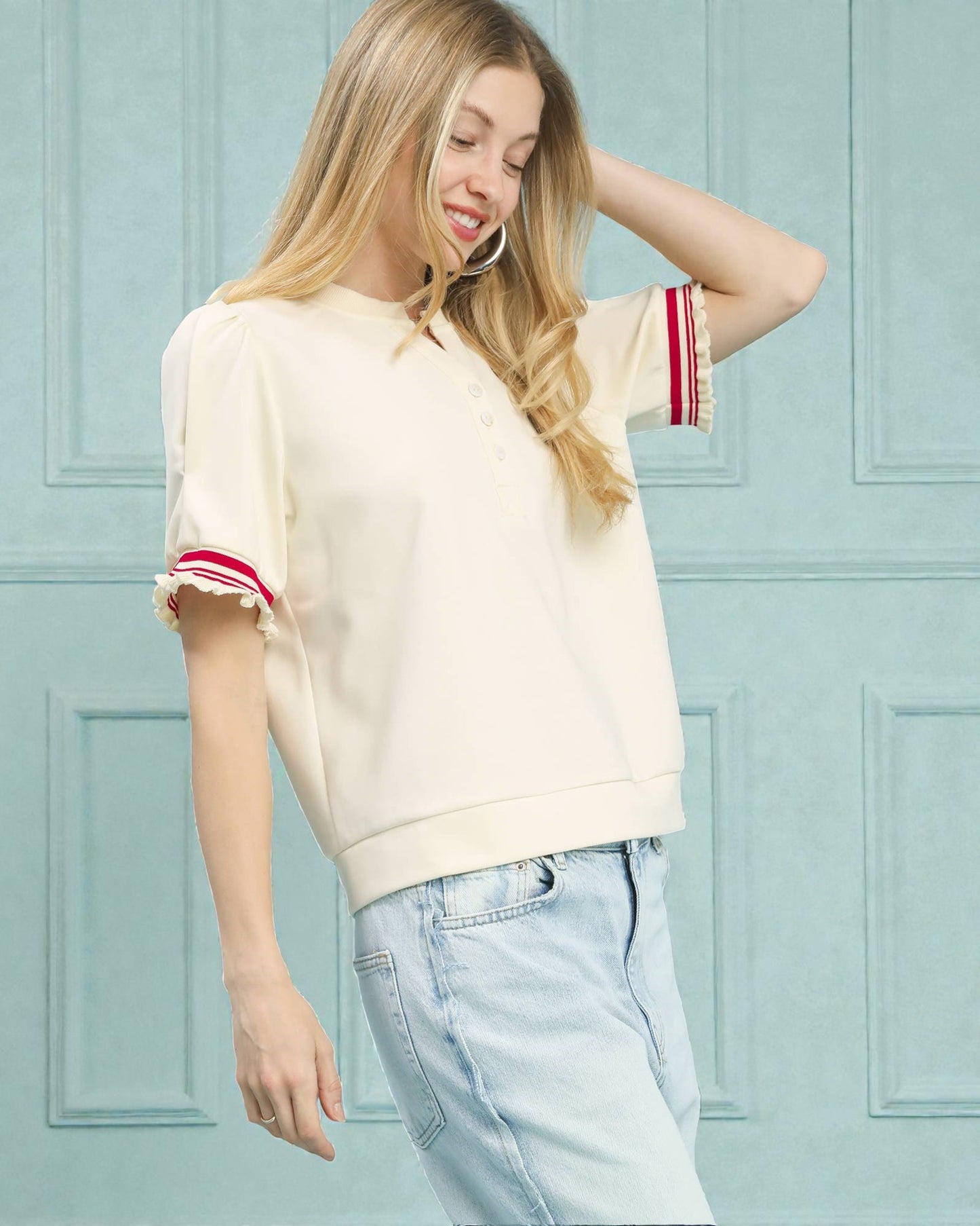 Woman wearing a cream-colored top with red and white trim and light blue jeans against a light blue paneled wall.