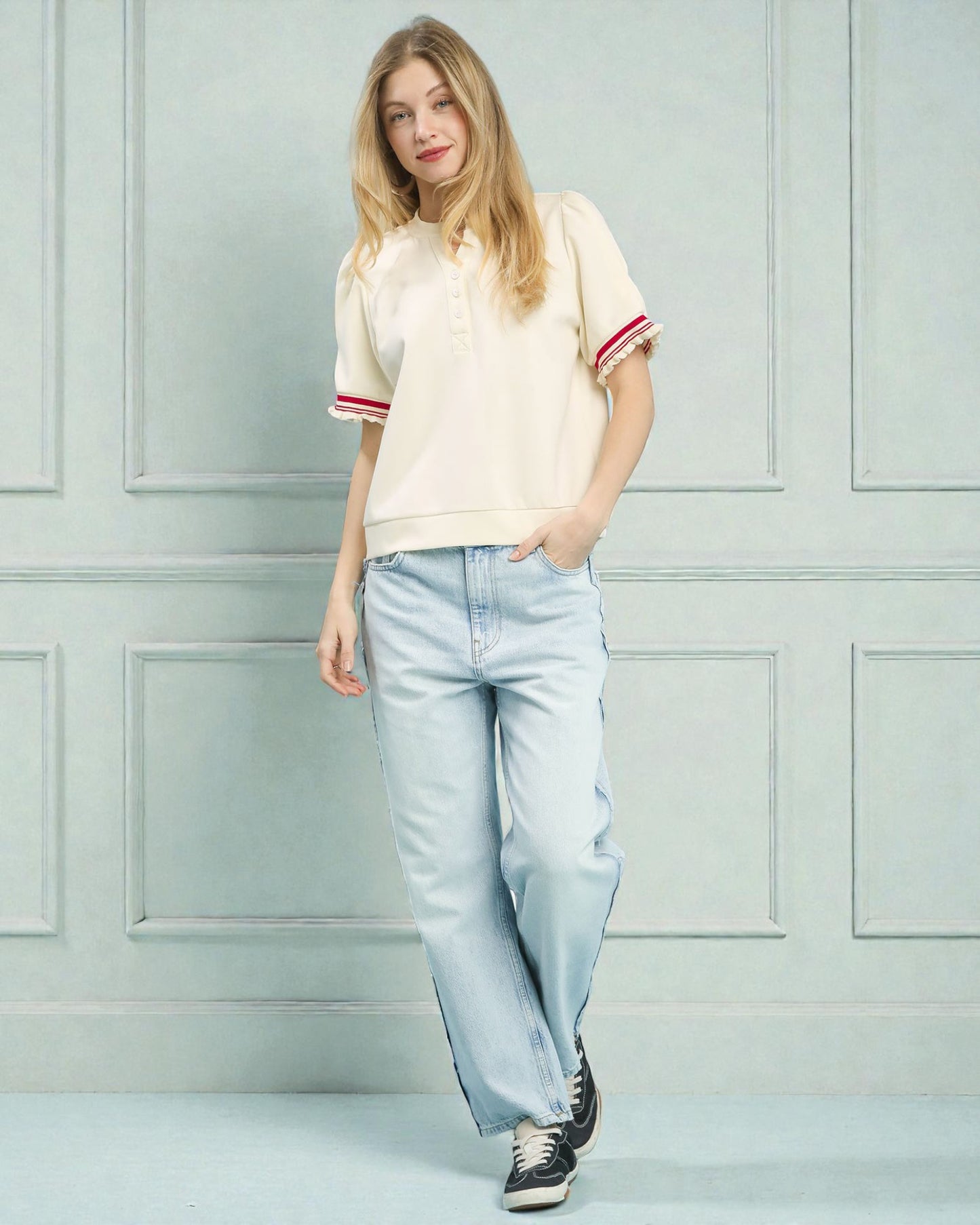 Woman wearing a cream-colored t-shirt with red trim and light blue jeans against a light gray wall.