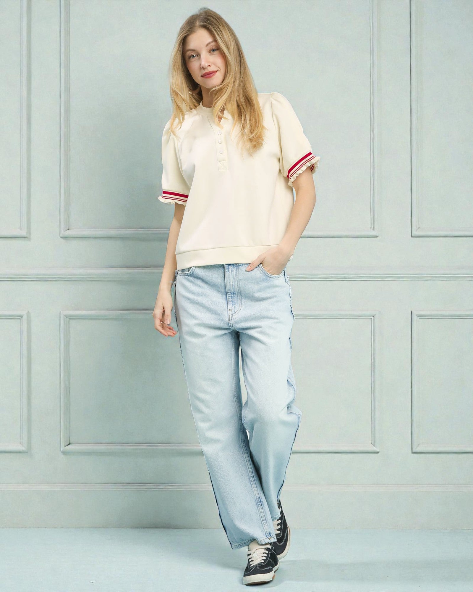 Woman wearing a cream-colored t-shirt with red trim and light blue jeans against a light gray wall.