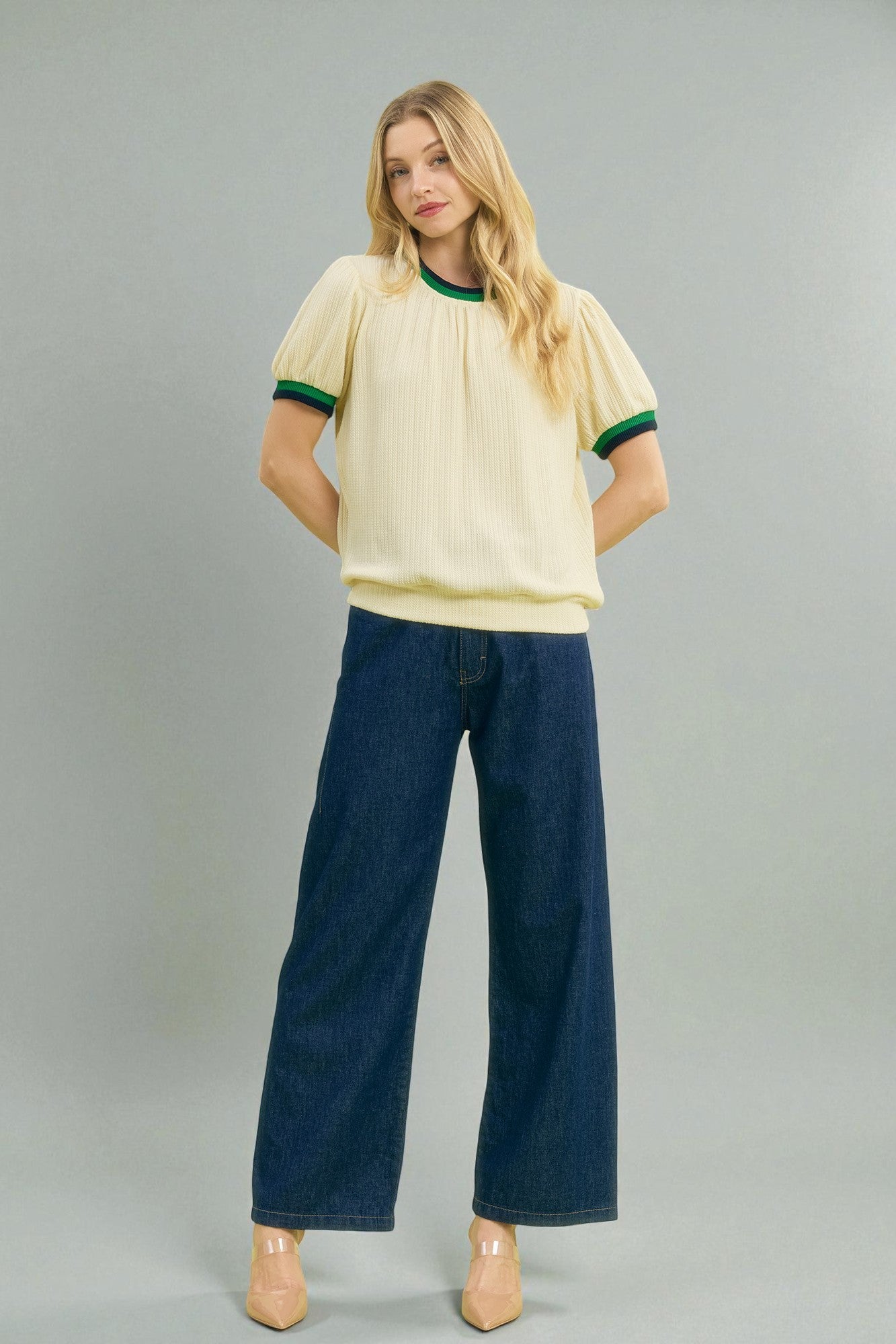 Woman wearing a yellow blouse with green trim and blue jeans on a gray background