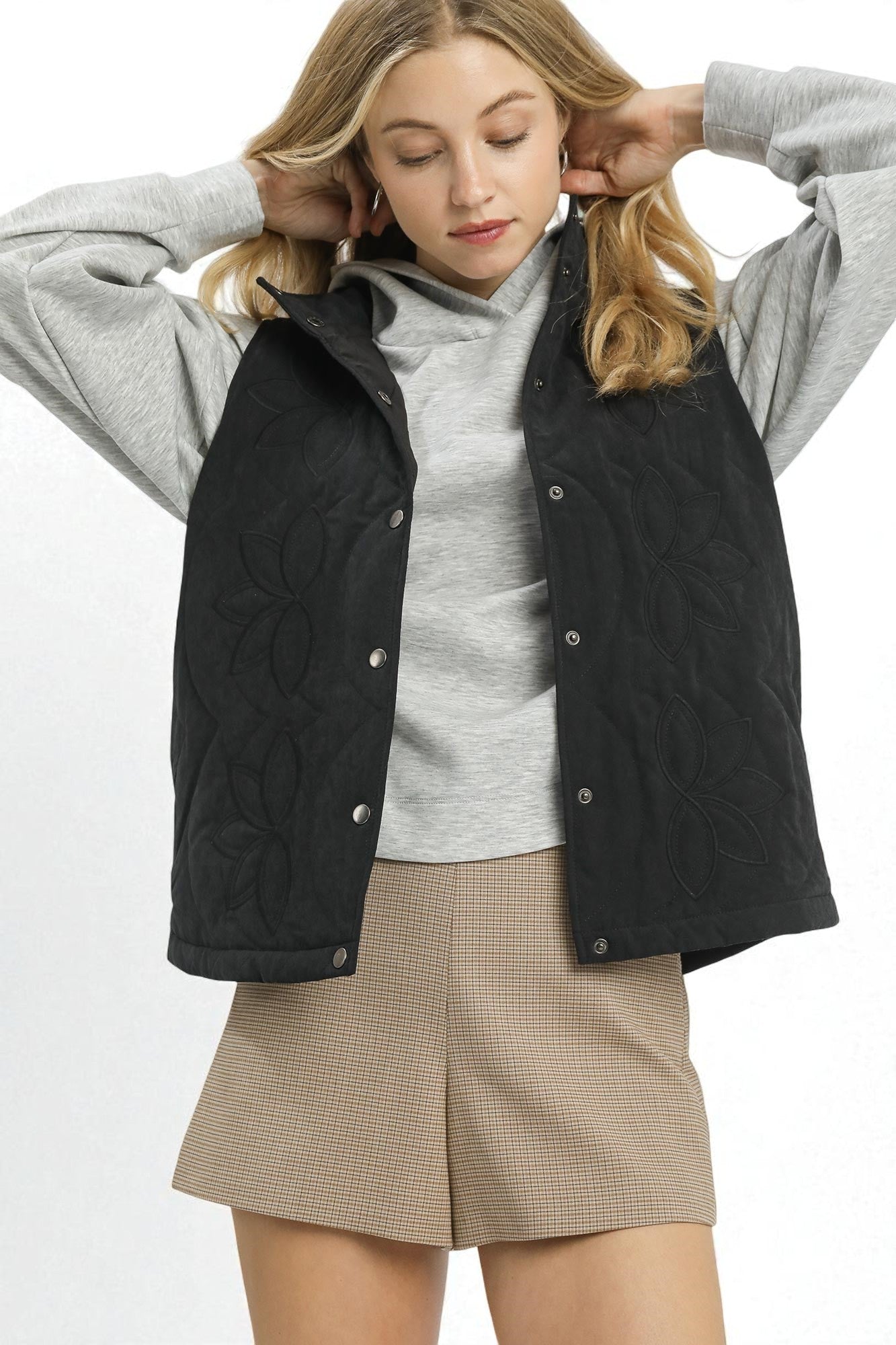 Woman wearing a black quilted vest over a gray sweater with beige shorts on a white background