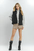 Woman wearing a black vest, gray shirt, beige shorts, and black boots on a white background