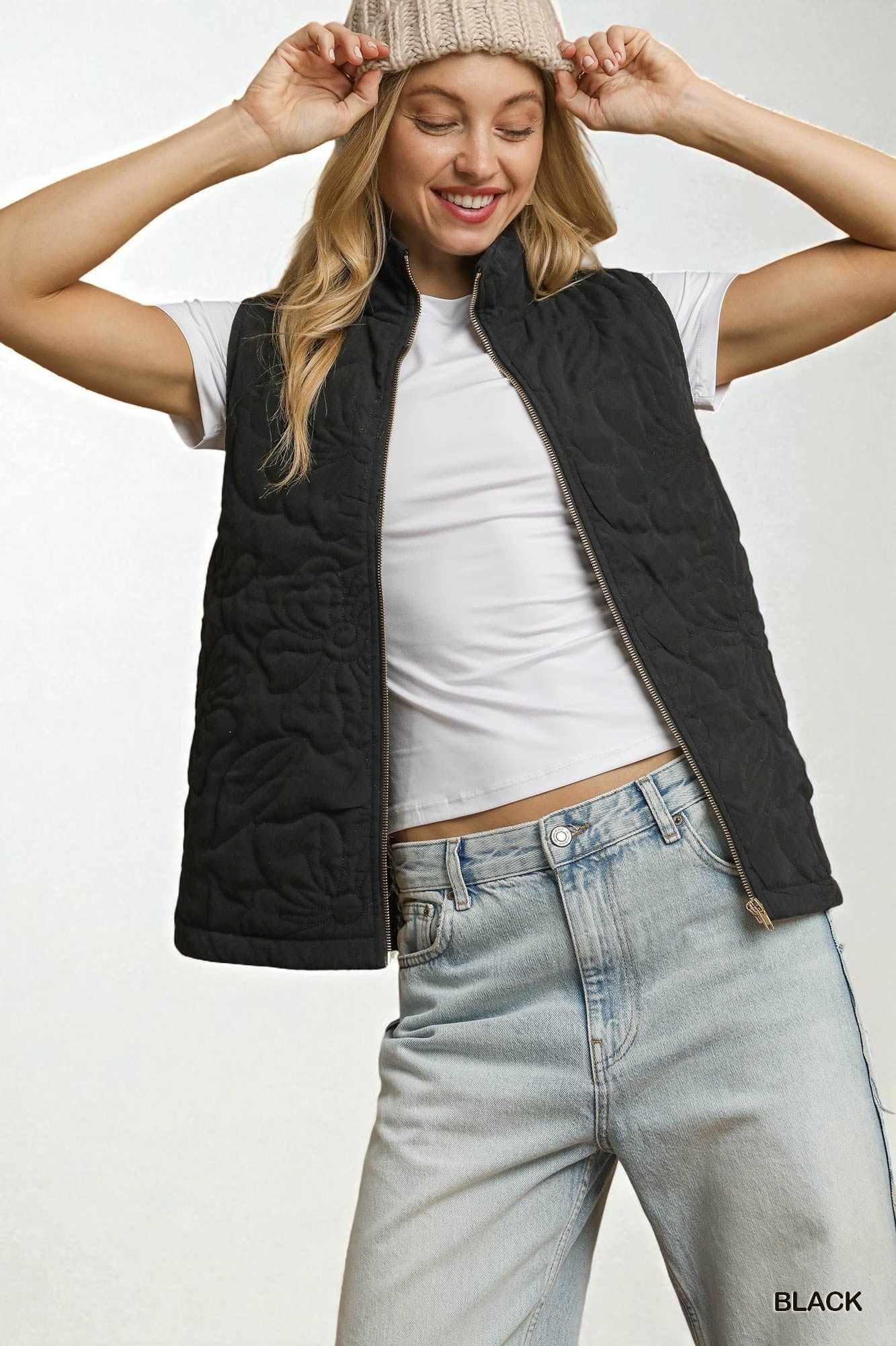 Woman wearing a black vest, white shirt, and light blue jeans on a plain background